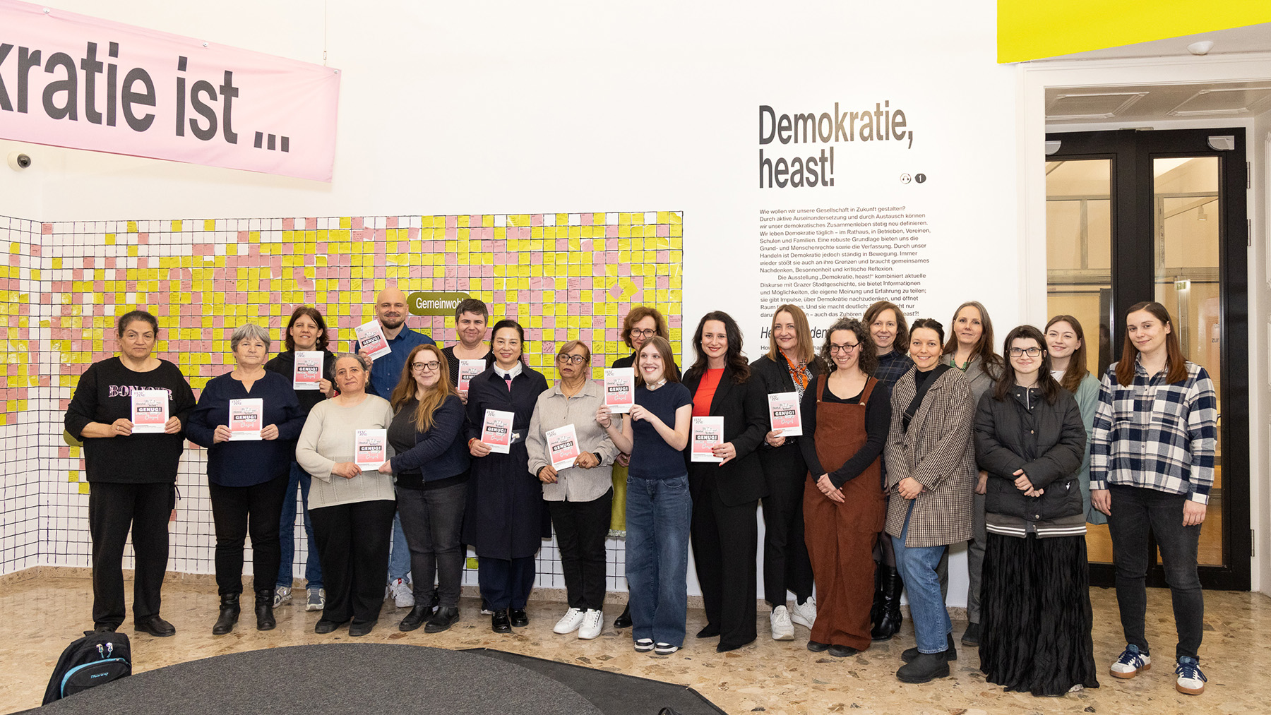 Eine Gruppe steht in einem Innenraum, viele mit Büchern in der Hand, vor einer Stellwand zum Thema Demokratie.