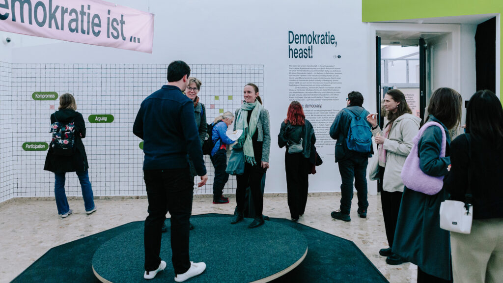 Menschen unterhalten sich in einem modernen Ausstellungsraum, an dessen Wand Demokratie, heast! prangt.