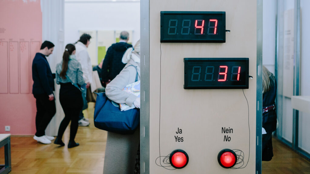 Die digitale Wahlanzeige zeigt "Ja" mit 47 Stimmen, "Nein" mit 31 Stimmen; dahinter Demokratie in Aktion.