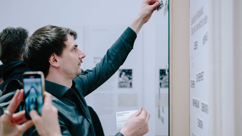 Ein Mann klebt ein Demokratie-Etikett auf einen Wandaufsteller, während eine andere Person ein Foto mit einem Telefon macht.