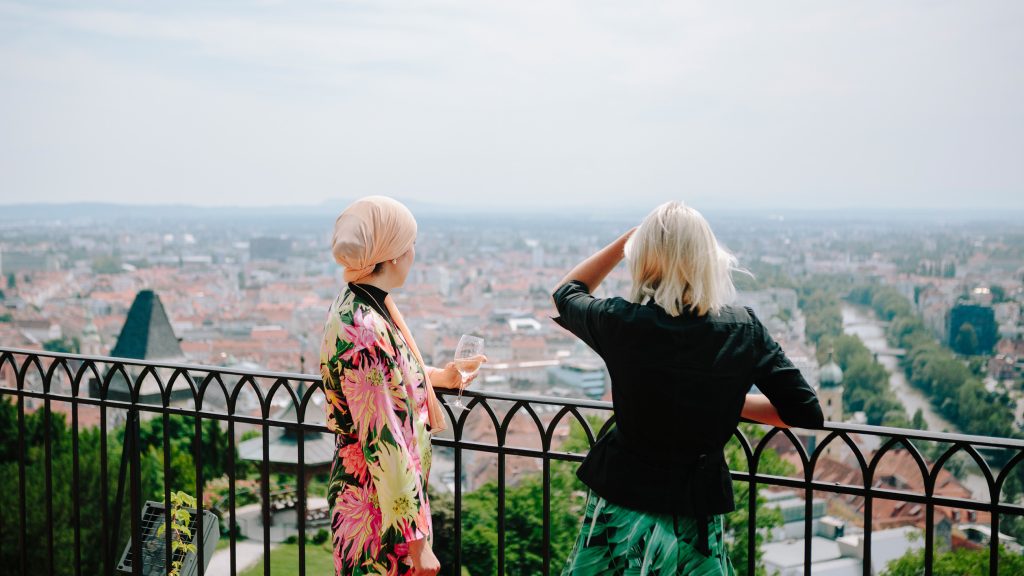 Zwei Frauen stehen an einem Geländer mit Blick auf eine Stadtlandschaft, eine hält ein Getränk in der Hand, beide schauen weg.