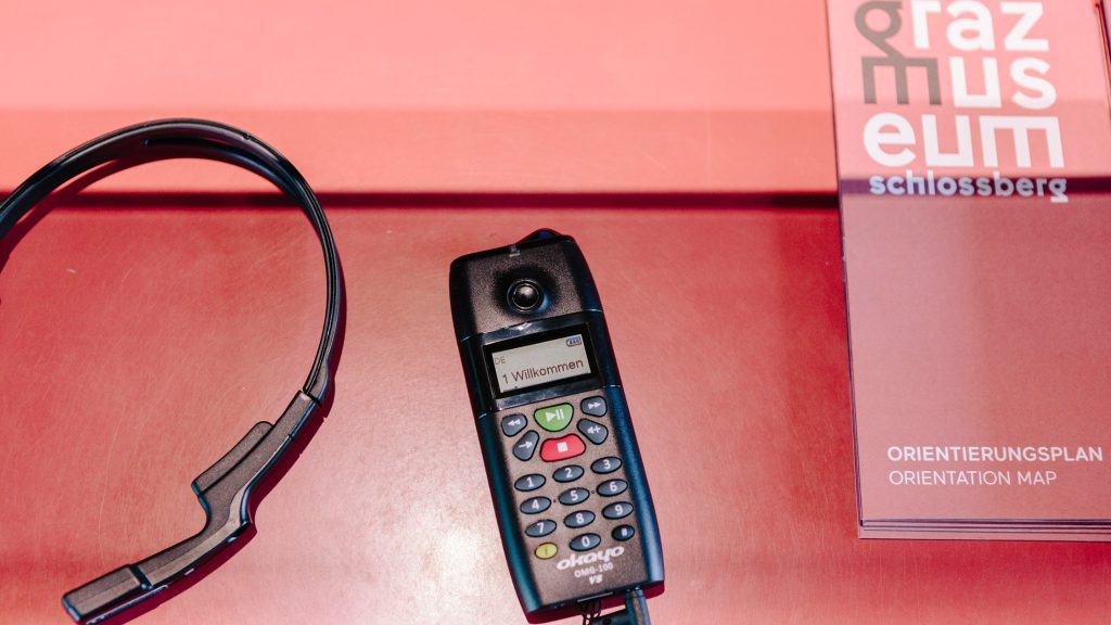Headset, barrierefreies Audioguide-Gerät und Orientierungskarte auf einer roten Fläche im Graz Museum.