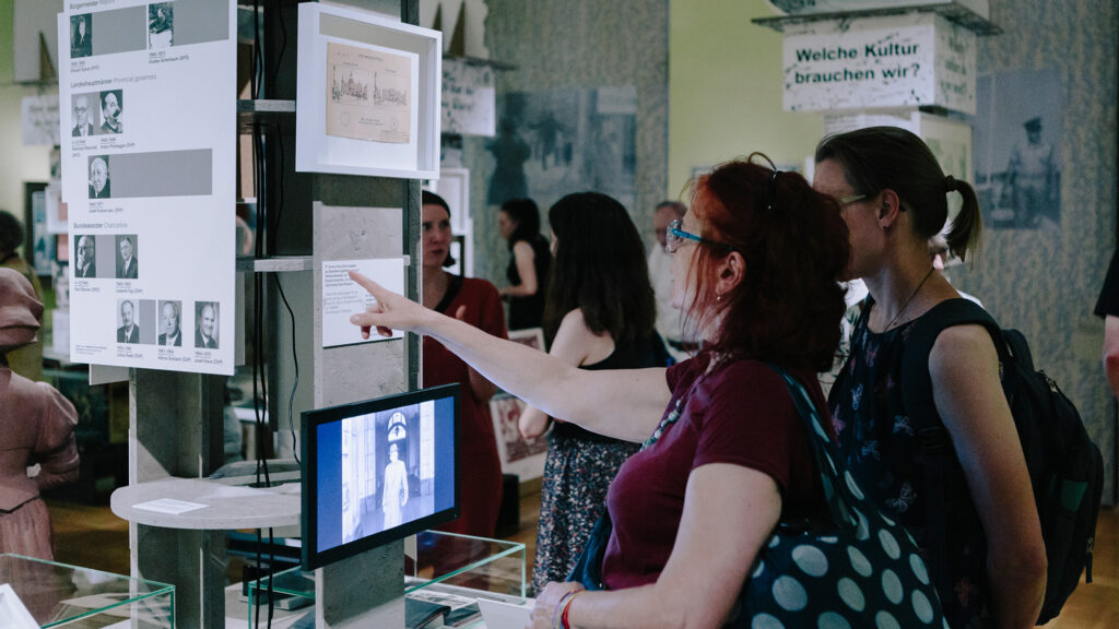 Menschen diskutieren über "Ins Ungewisse"-Ausstellungen in einem Grazer Museum mit Fotos, Text und digitaler Darstellung.
