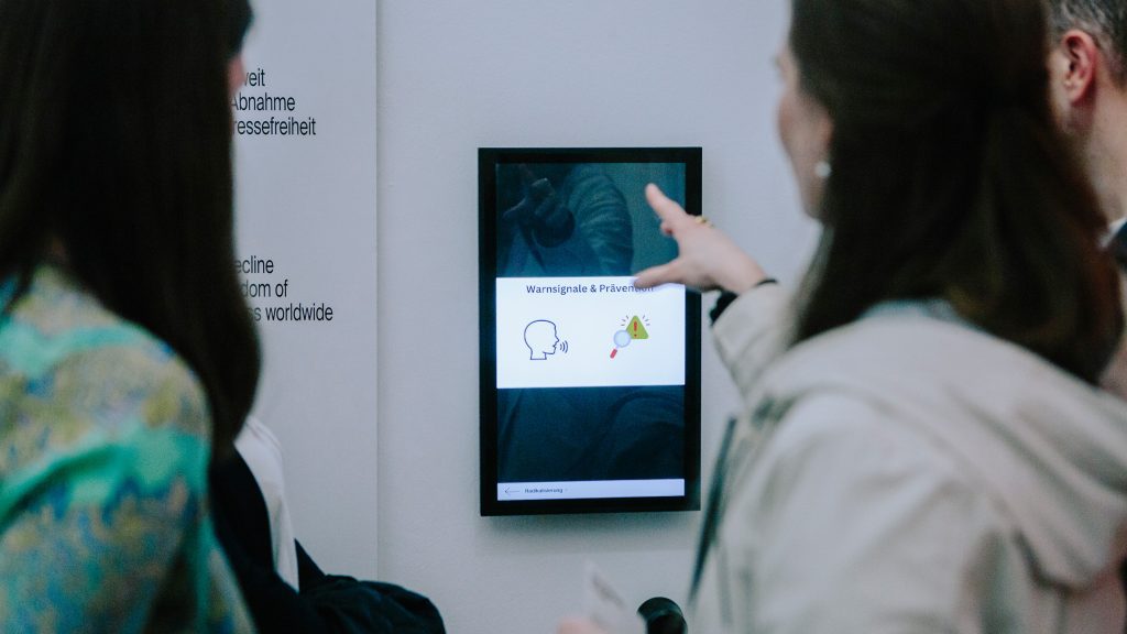 Person zeigt auf einen an der Wand montierten Bildschirm zum Thema Demokratie, während andere in der Nähe stehen und mitmachen.