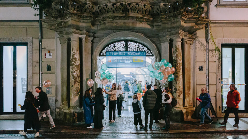 Bei der Langen Nacht der Museen versammeln sich Menschen mit Luftballons vor dem kunstvollen Torbogen des Graz Museums.