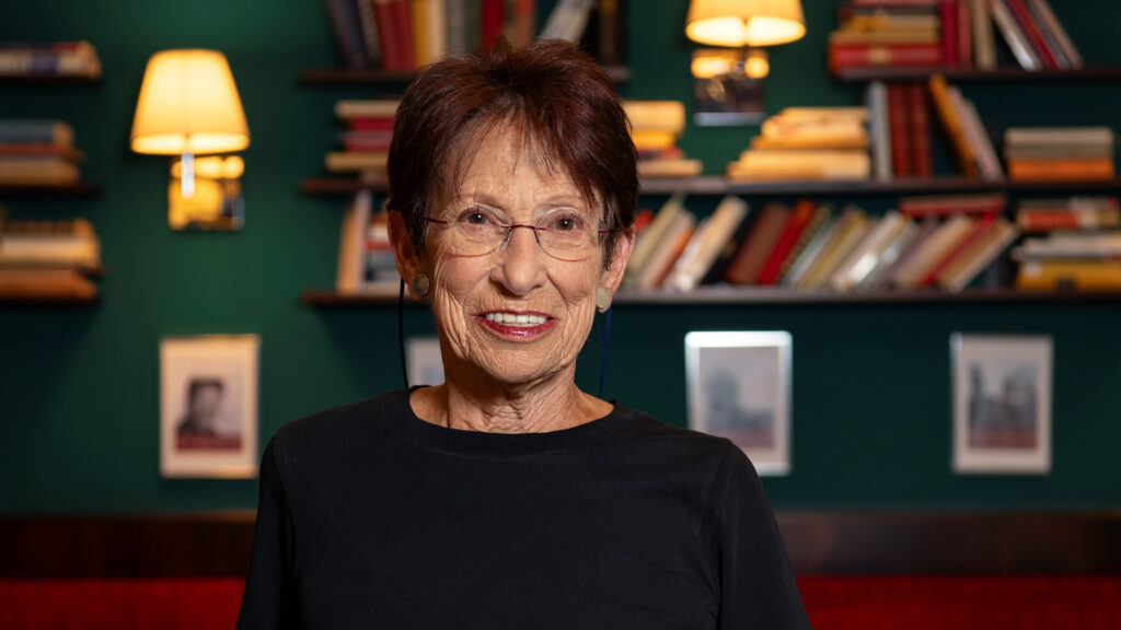 Eine ältere Frau mit Brille steht lächelnd vor einem Bücherregal und recherchiert über Desinformation.