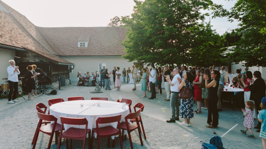 Freund*innen versammeln sich im Freien an runden Tischen und genießen eine Live-Band auf der Bühne des Graz Museums.