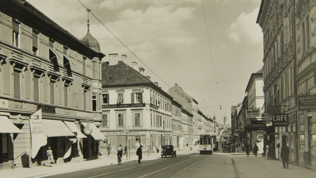 Schwarz-Weiß-Foto einer Grazer Stadtstraße mit Fußgängern, Autos und einer Straßenbahn, 1938-1945.