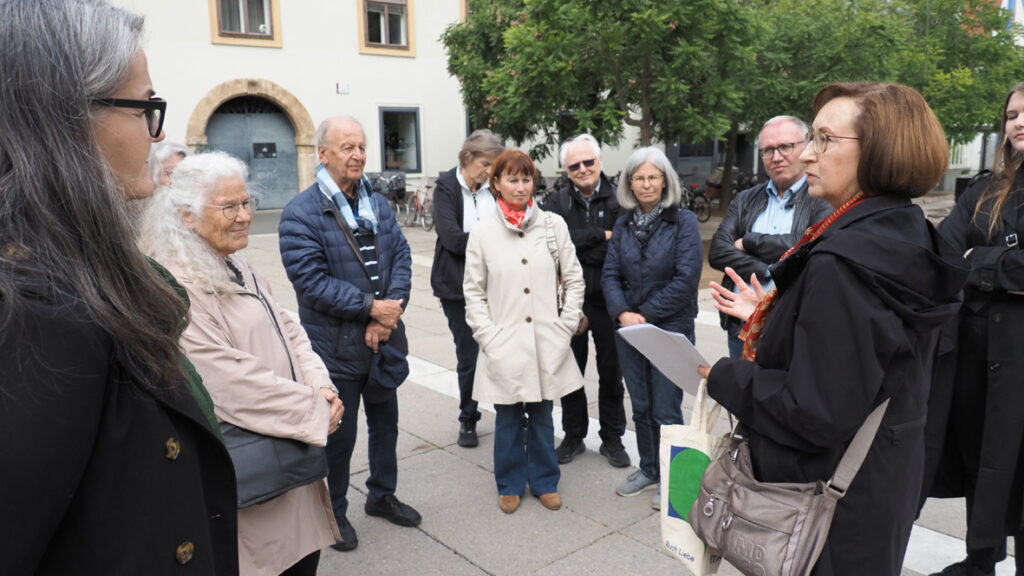 Eine Gruppe von Menschen in Graz hört im Freien zu, als eine Frau vom Graz Museum spricht und Papiere hält.