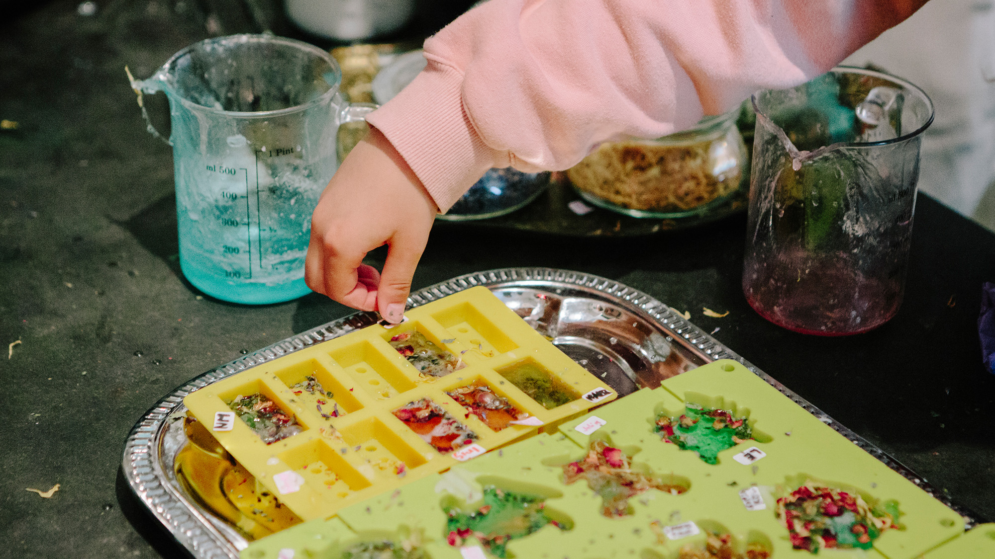 Eine Person fertigt Kunsthandwerk in Silikonformen auf einem Tablett an, Messbecher stehen daneben.