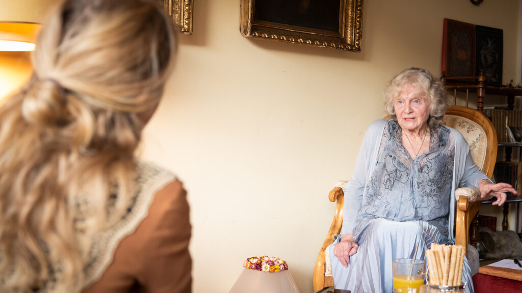 Eine ältere Frau schwelgt in Erinnerungen an die verlorene Kindheit, während sie sich in einem gemütlichen Zimmer mit einer jüngeren Frau unterhält.
