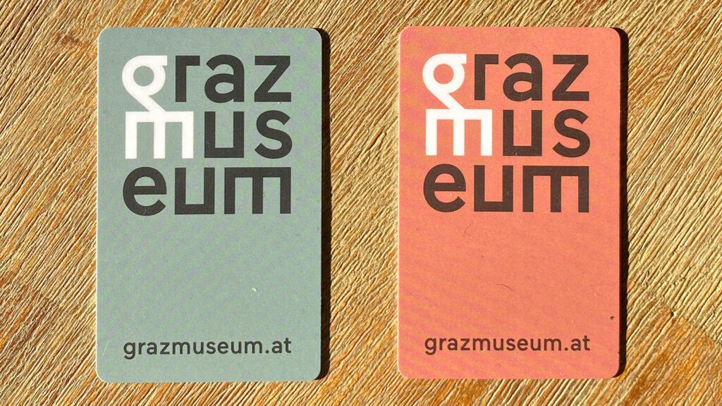 Zwei rechteckige Karten mit Text und Website des grazmuseums, eine grau und eine rot, auf einer Holzoberfläche.