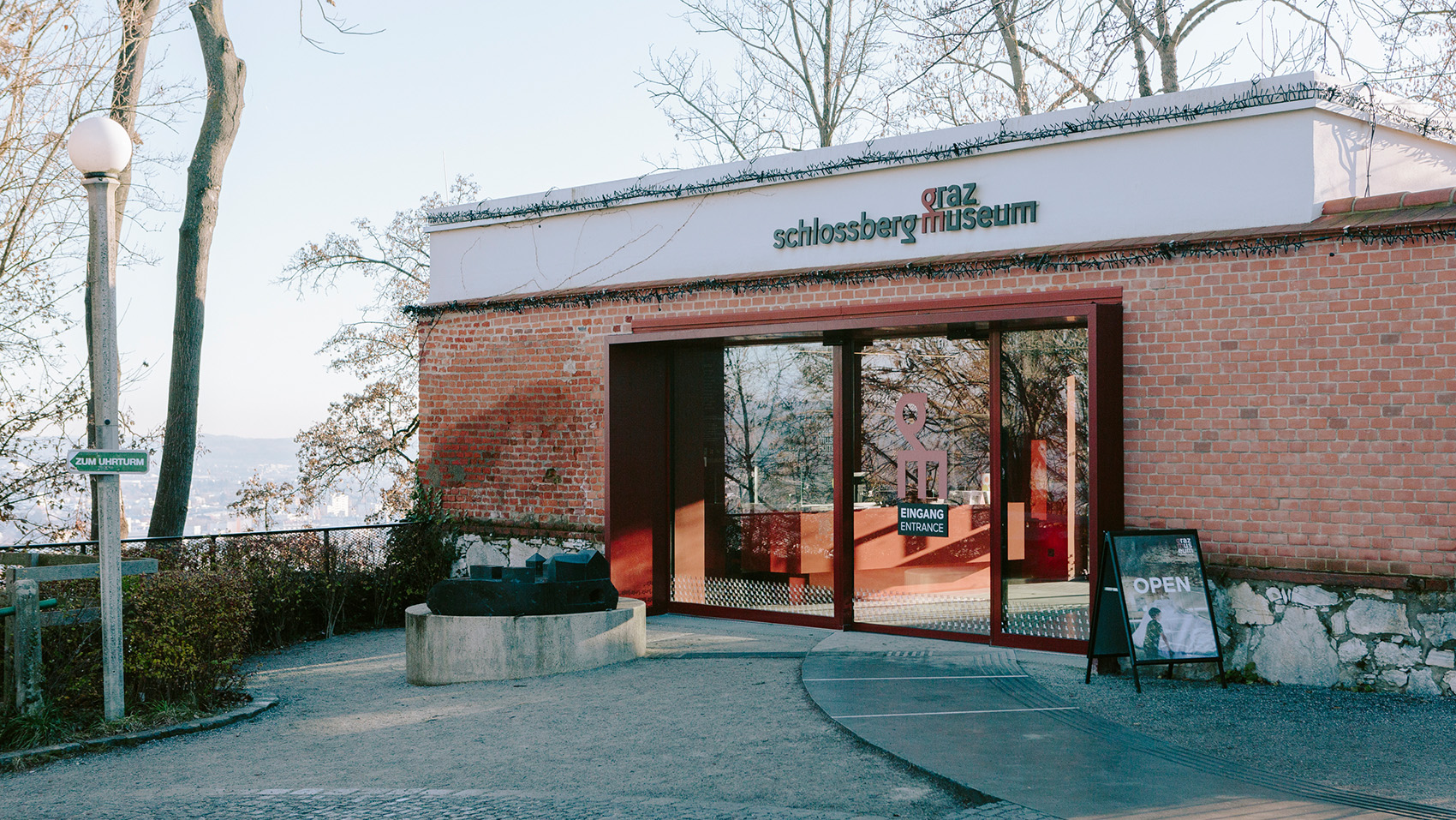 Eingang zum Grazer Schlossbergmuseum mit Backsteinfassade, roter Tür und offenem Schild.
