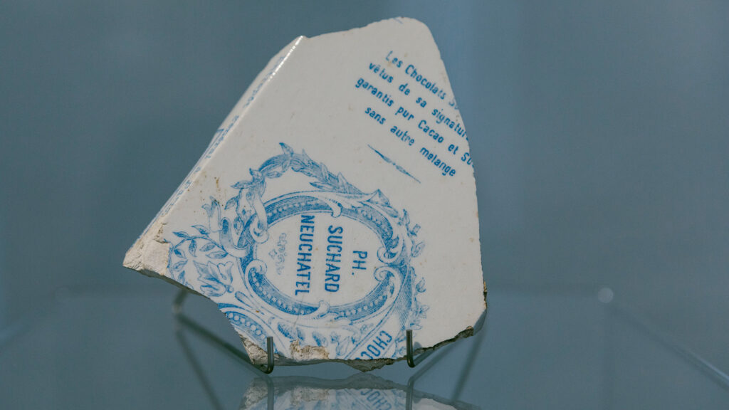 Ein zerbrochener weißer Keramikscherben mit blauer französischer Aufschrift, ausgegraben im Lager Liebenau, auf Ausstellungsständern.