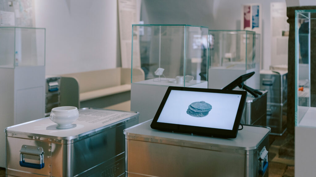 Ausstellungsraum eines Museums mit Vitrinen und einem Tablet, auf dem das Bild eines Hamburgers zu sehen ist, „Erinnern“.
