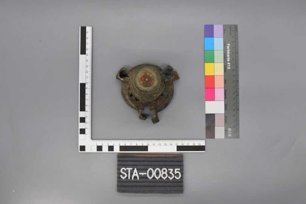 Metallartefakt mit vier Vorsprüngen, vermessen und mit STA-00835 gekennzeichnet, mit Farb- und Maßstabsbalken.