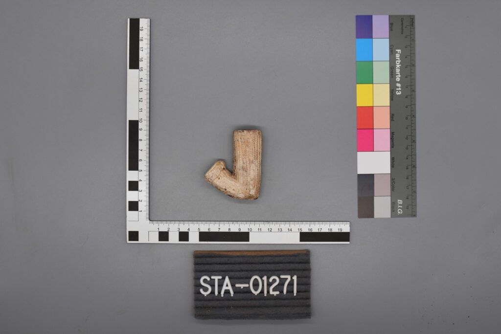Ein kleines, L-förmiges Artefakt neben Messwerkzeugen und einem Etikett mit der Aufschrift STA-01271.