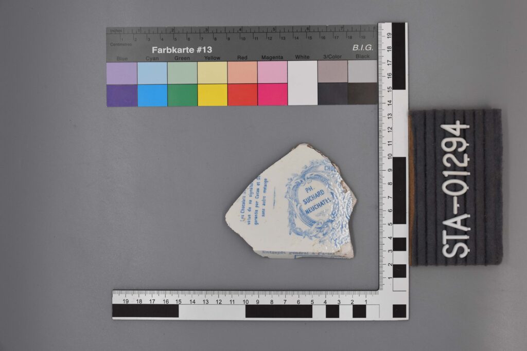 Ein zerbrochenes Keramikstück mit blauem Text und Emblem, dargestellt mit Maß- und Farbskalen.