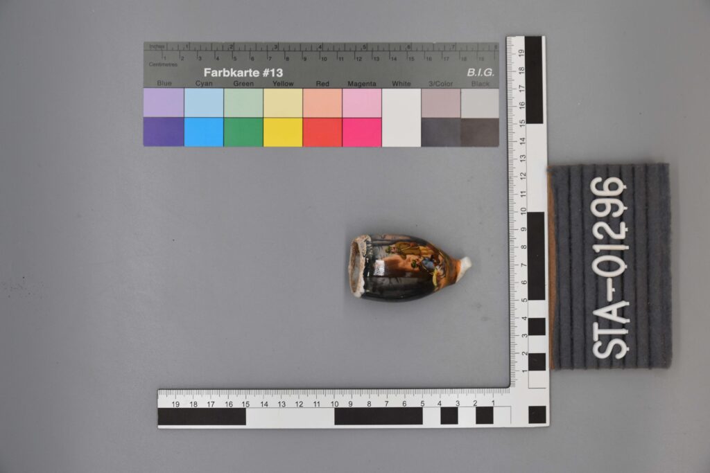 Kleines bemaltes Keramikfragment neben einer Messskala, einer Farbkarte und dem Etikett STA-01296.