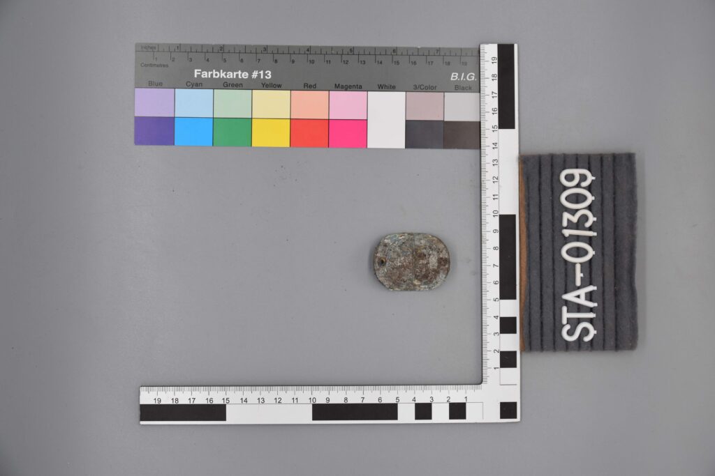 Ein kleiner, korrodierter Metallgegenstand neben einer Messskala mit der Aufschrift STA-01309.