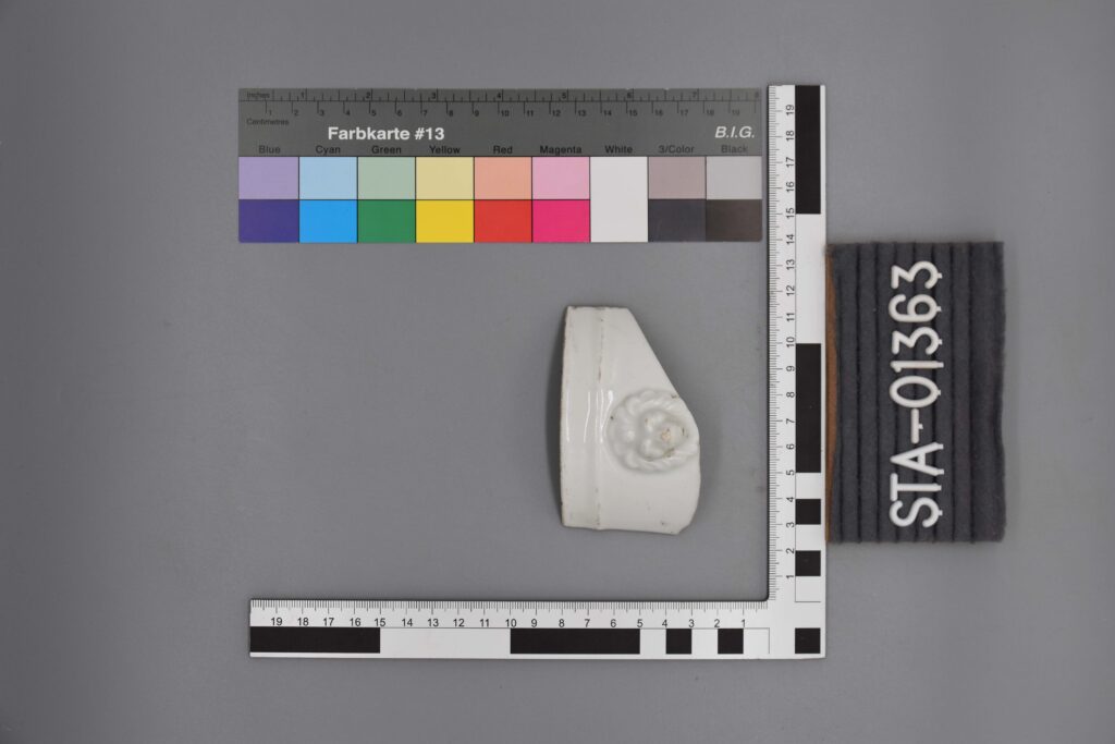 Weißes Keramikfragment mit floralem Muster, daneben Messskalen und Etikett STA-01363.