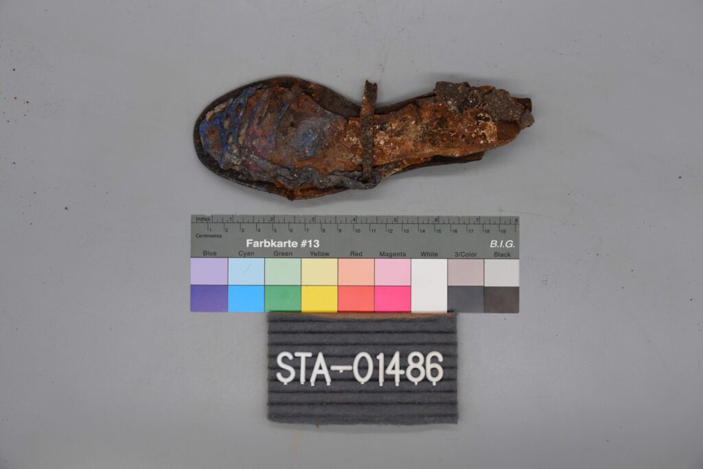 Ein verrosteter, verrotteter Schuh über einer Farbkarte und einem Etikett mit der Aufschrift STA-01486 auf einer grauen Fläche.