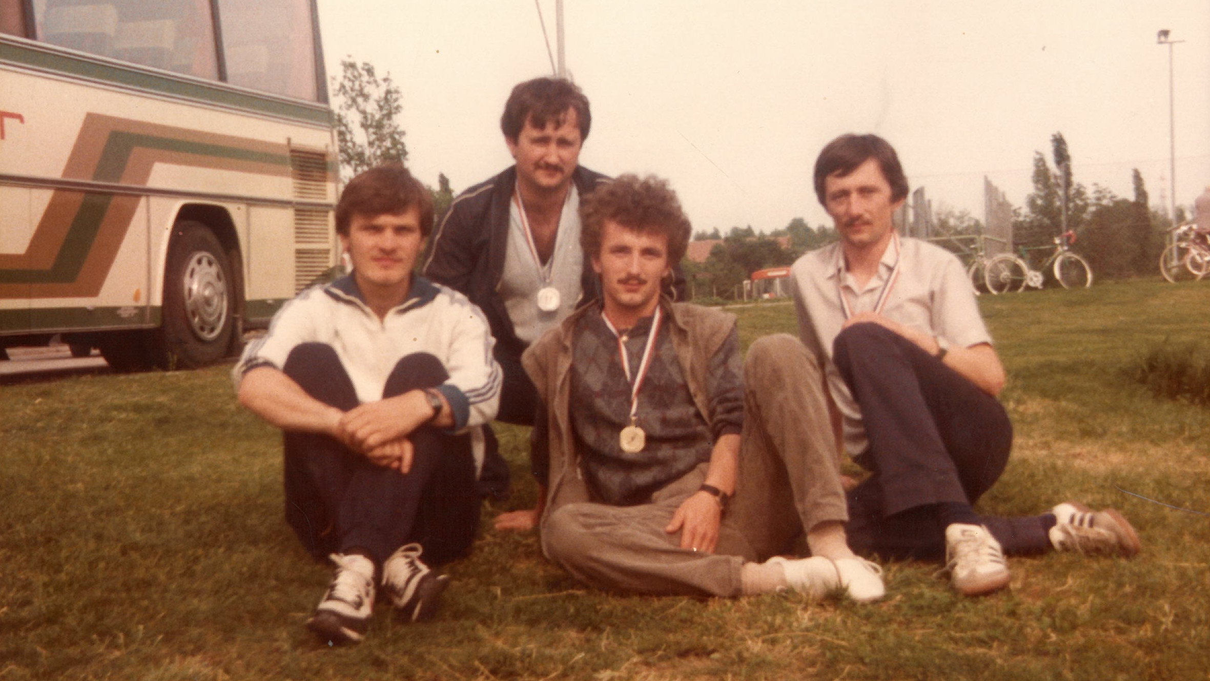 Vier Männer mit Medaillen sitzen im Gras neben einem Bus, ihre Zukunft ist geprägt von Migrationsgeschichte.