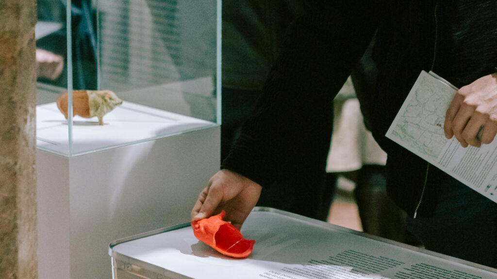 Person legt roten Gegenstand in die Museumsauslage, Karte in der Hand - Teil der Sonderführung im Lager Liebenau.