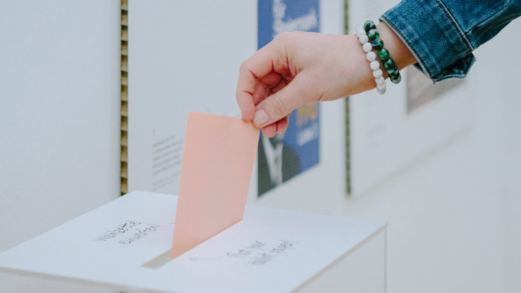 Eine Person wirft bei der Gemeinderatswahl einen rosa Stimmzettel in eine weiße Wahlurne.