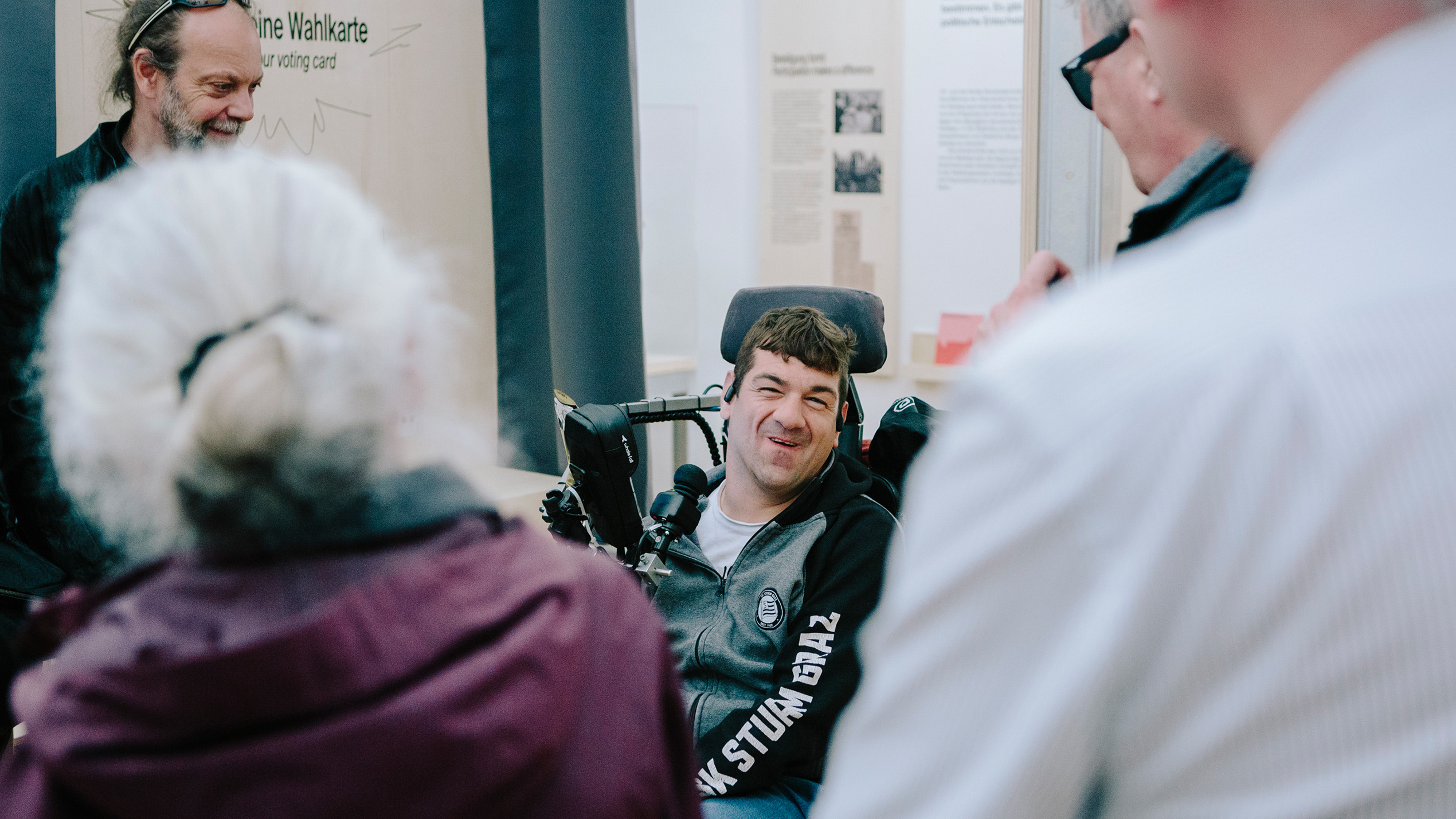 Ein Mann im Rollstuhl lächelt, während er mit einer Gruppe im Graz Museum über Inklusive Politik diskutiert.