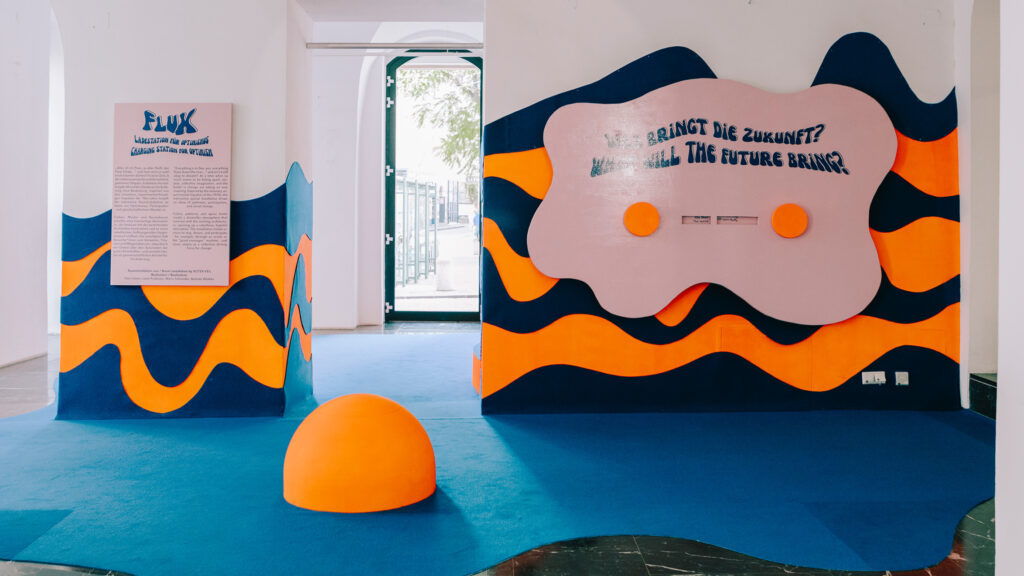 Heller Ausstellungsraum mit wellenförmigen blauen Wänden im Flux-Stil und einer orangefarbenen Kuppel auf blauem Boden.
