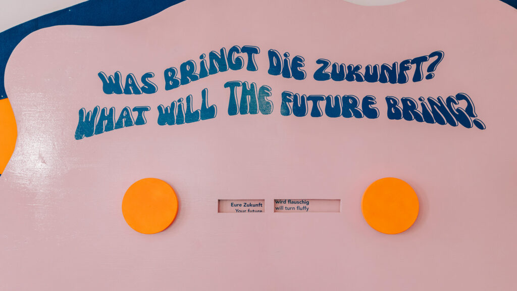 Auf einer rosa Wand, die sich ständig verändert, steht auf Deutsch und Englisch: „Was wird die Zukunft bringen?“.