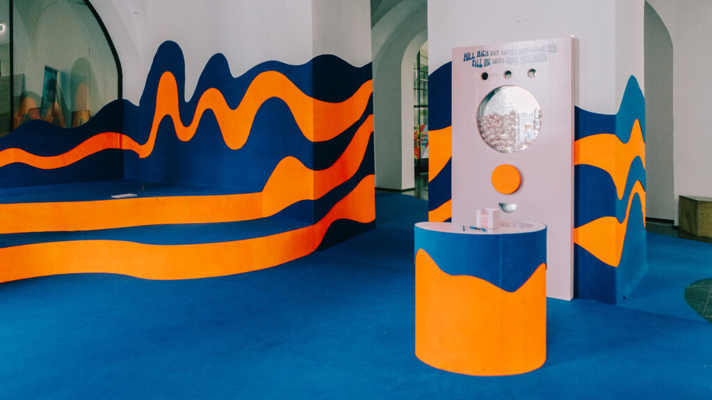 Raum mit blauer und orangefarbener „Flux“-Wandgestaltung, einer Spiegelinstallation und einem Sockel auf blauem Boden.