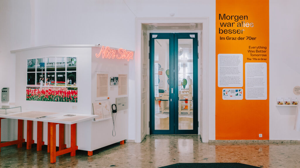 Museum exhibit with ‘70s in Graz text panels, neon sign, and tulip photo by double glass doors.
