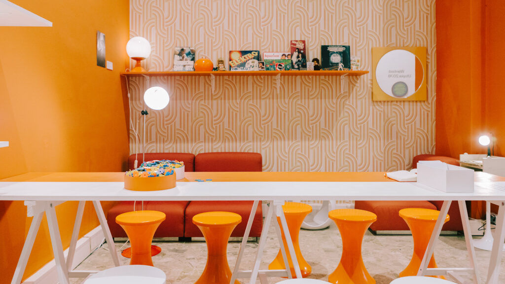 Modern room with orange walls, ‘70s in Graz vibe, orange stools, white table, and shelf decor.