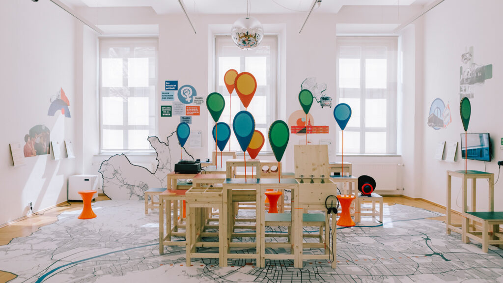 Modern exhibition room with colorful markers, wooden furniture, and a ‘70s in Graz map on the floor.