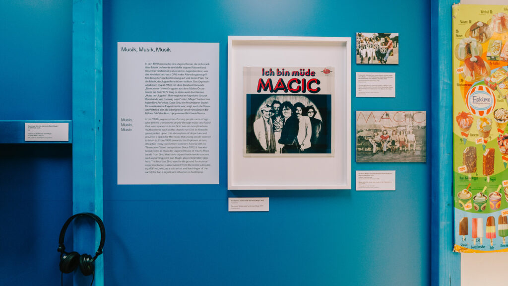 Exhibit wall with framed album cover, text, photos, headphones—celebrating 70s in Graz.