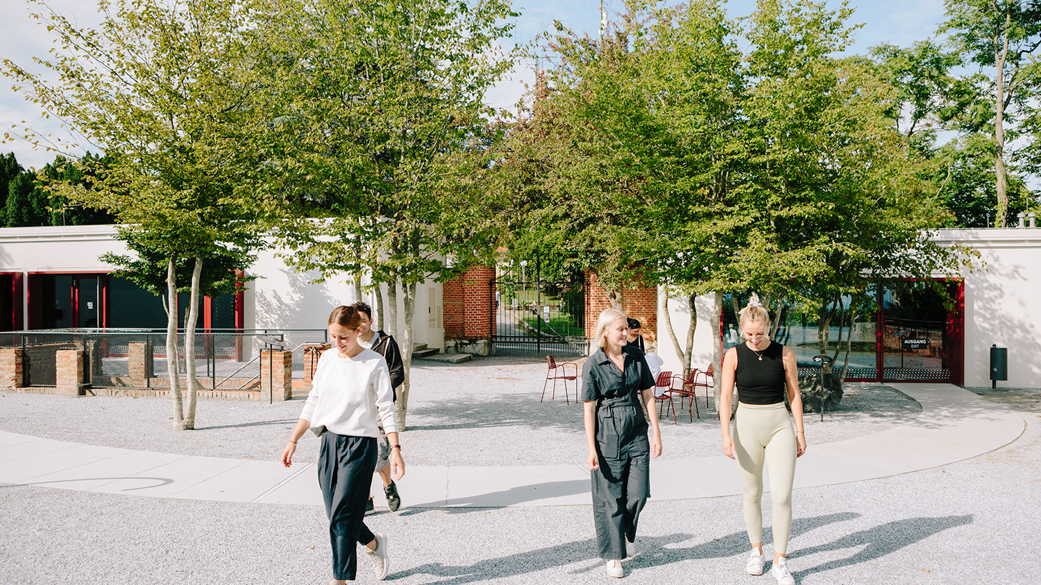 Vier Personen genießen einen sonnigen Rundgang in der Nähe eines Gebäudes mit Bäumen und Sitzgelegenheiten im Freien.