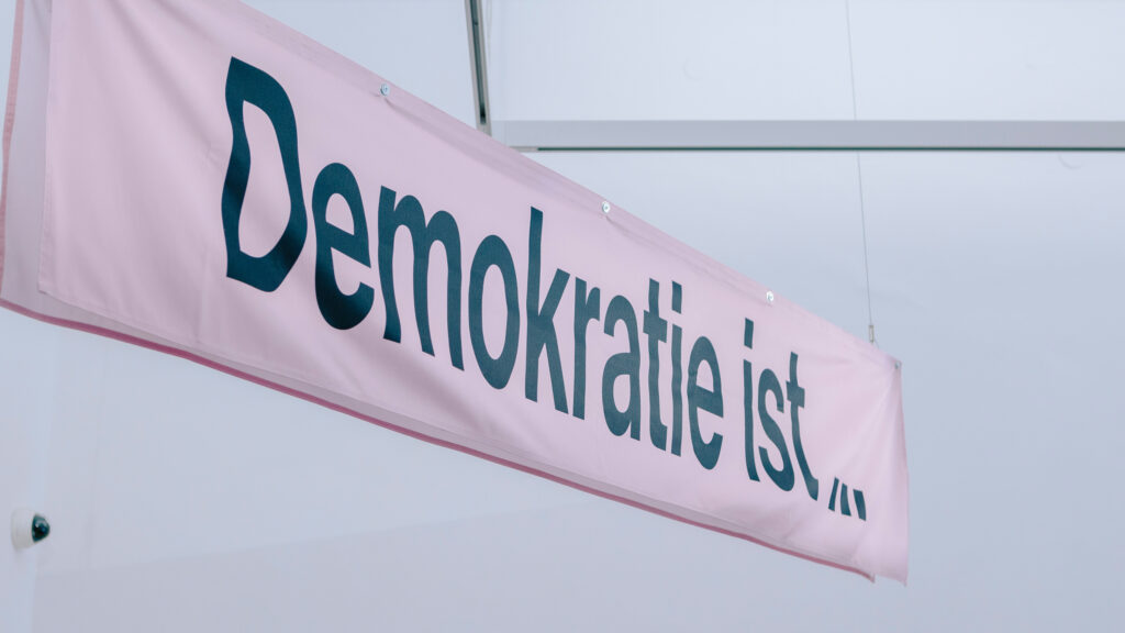 Rosa Banner mit fetter schwarzer Schrift „Demokratie ist...“, das für Vielfalt und Menschenrechte wirbt.