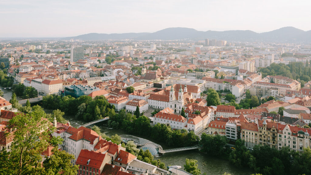 Luftaufnahme von Graz mit roten Dächern, dem Fluss, dem Schlossberg und den Hügeln in der Ferne unter klarem Himmel.