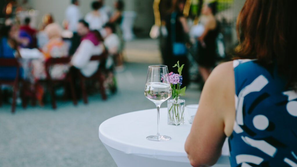 Eine Frau sitzt an einem Cocktailtisch im Graz Museum und nippt neben einem Strauß frischer Blumen an einem Glas Weißwein.