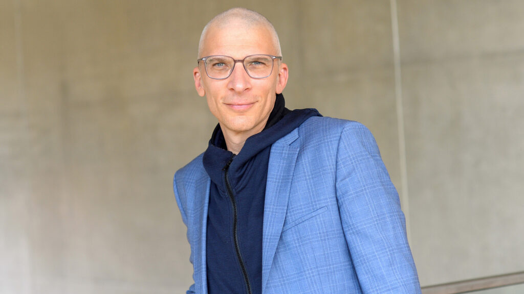 Eine Person mit offenen Augen, Brille und einem blauen Blazer steht vor einer hellen Wand.