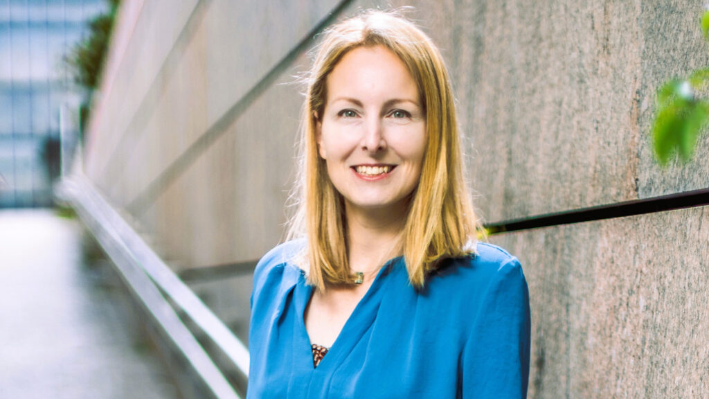 Eine Frau mit blonden Haaren in einem blauen Hemd steht lächelnd neben einer Steinmauer – „Demokratie schützen“.