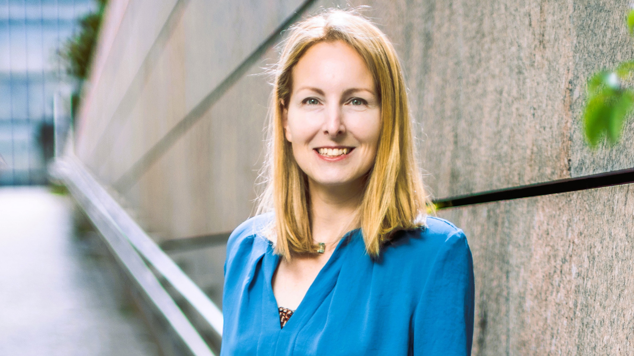 Eine Frau mit blonden Haaren in einem blauen Hemd steht lächelnd neben einer Steinmauer – „Demokratie schützen“.