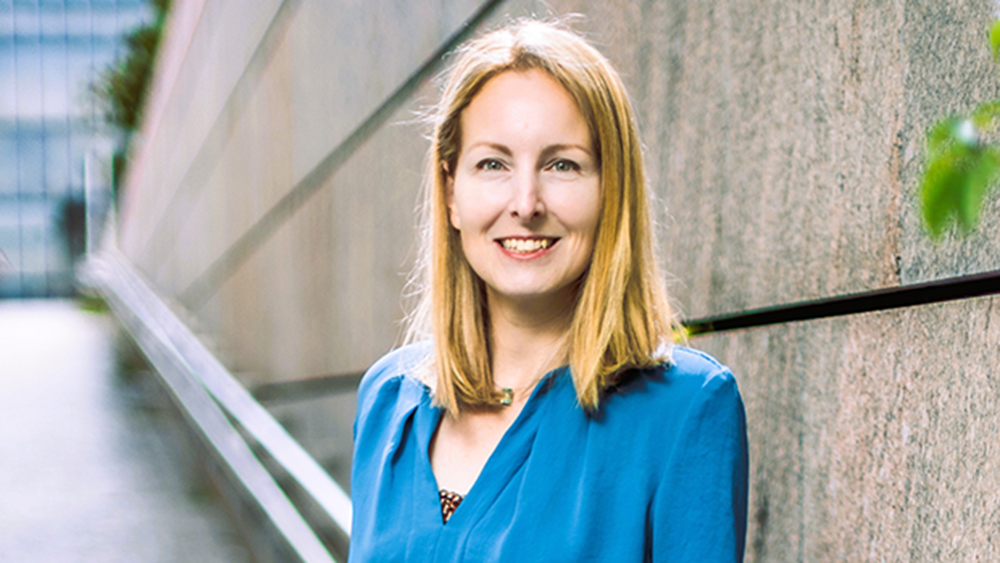 Eine Frau mit blonden Haaren in einer blauen Bluse steht lächelnd im Freien vor einer Steinmauer für die Demokratie.
