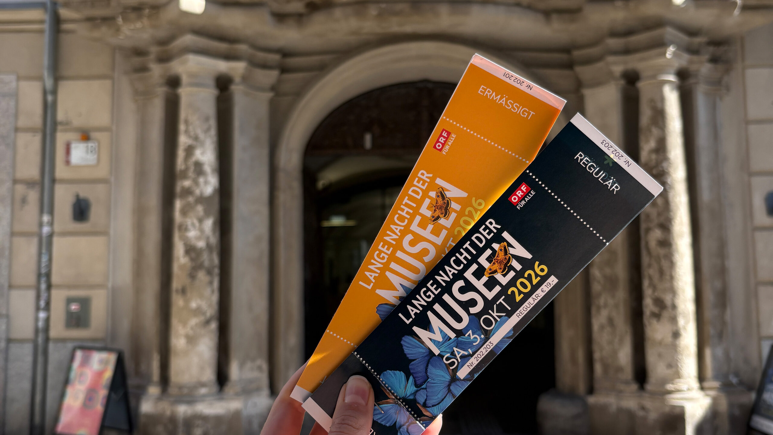 Vor dem Eingang eines alten Steingebäudes werden zwei Eintrittskarten für eine Museumsveranstaltung hochgehalten.